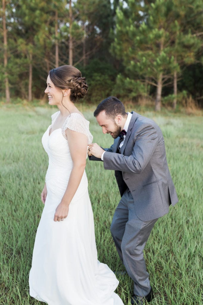 groom helping bride zip up the back of wedding dress in big field