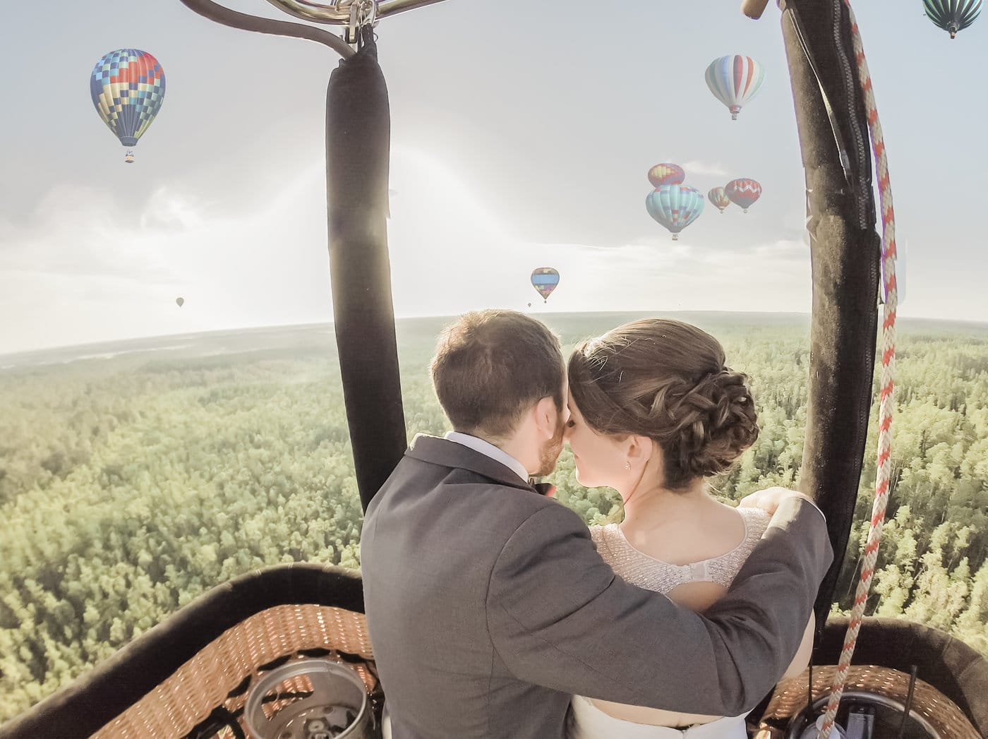 bride and groom pressing foreheads together inside basket of hot air balloon with balloons all around the horizon in the air