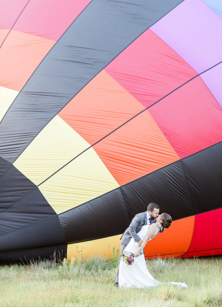 bride and groom kissing in front of colorful hot air balloon as it is being blown up
