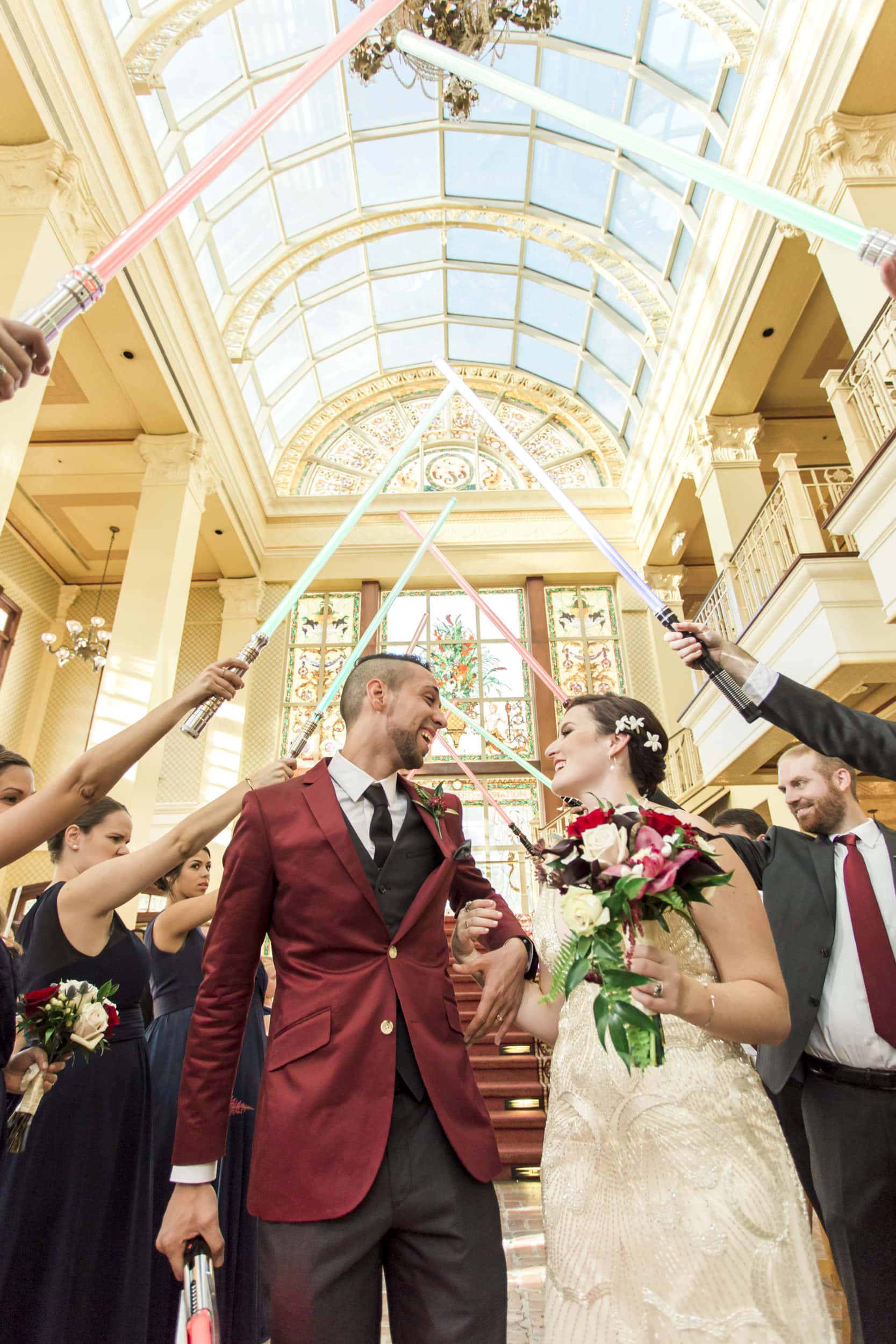 bride with white dress with red and white flowers with groom with red suit with wedding guests holding light sabers with large glass window ceiling in background at Star Wars Wedding