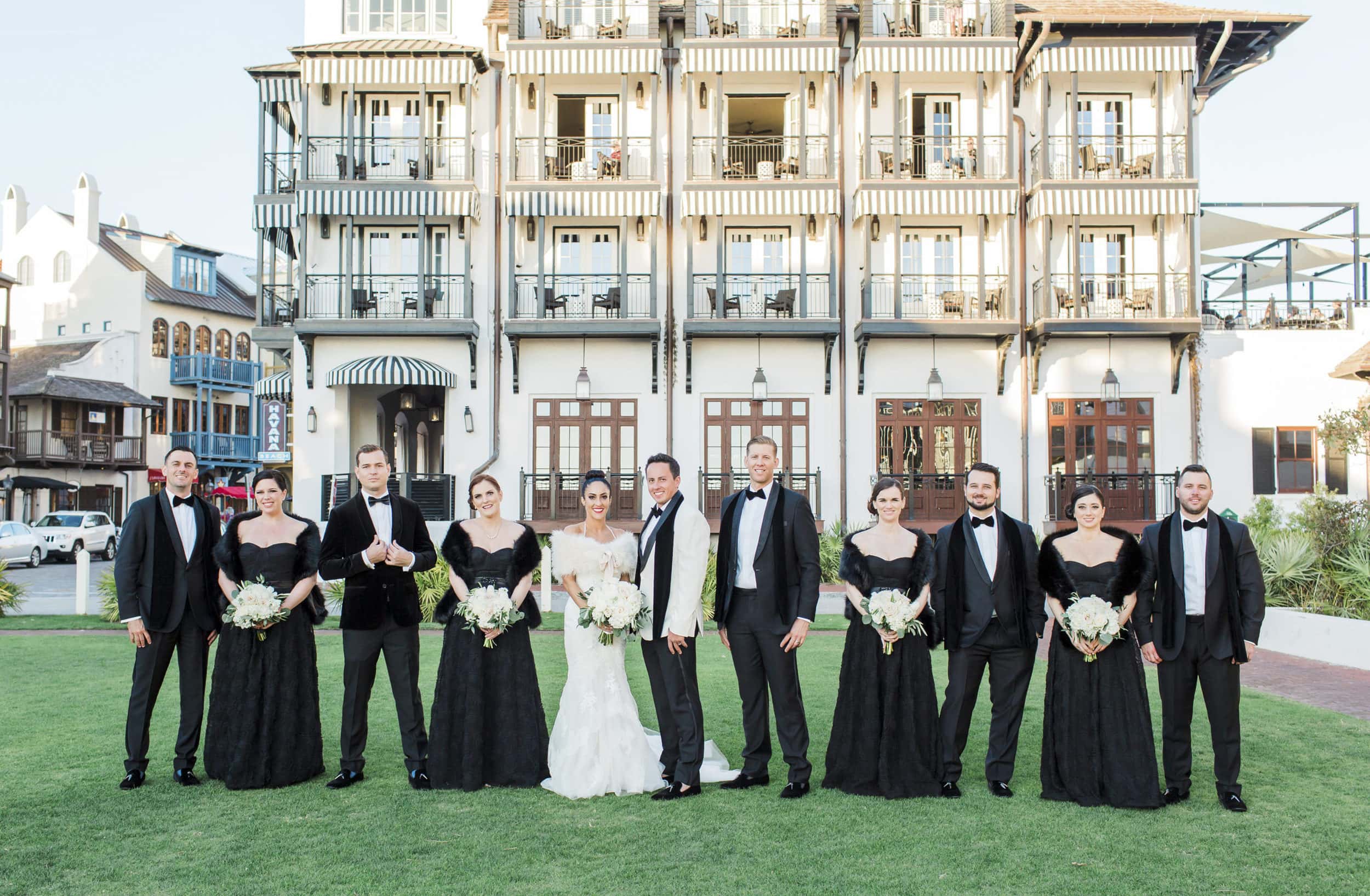 bride and groom standing on green grass in front of the pearl hotel in rosemary beach with bridesmaids and groomsmen
