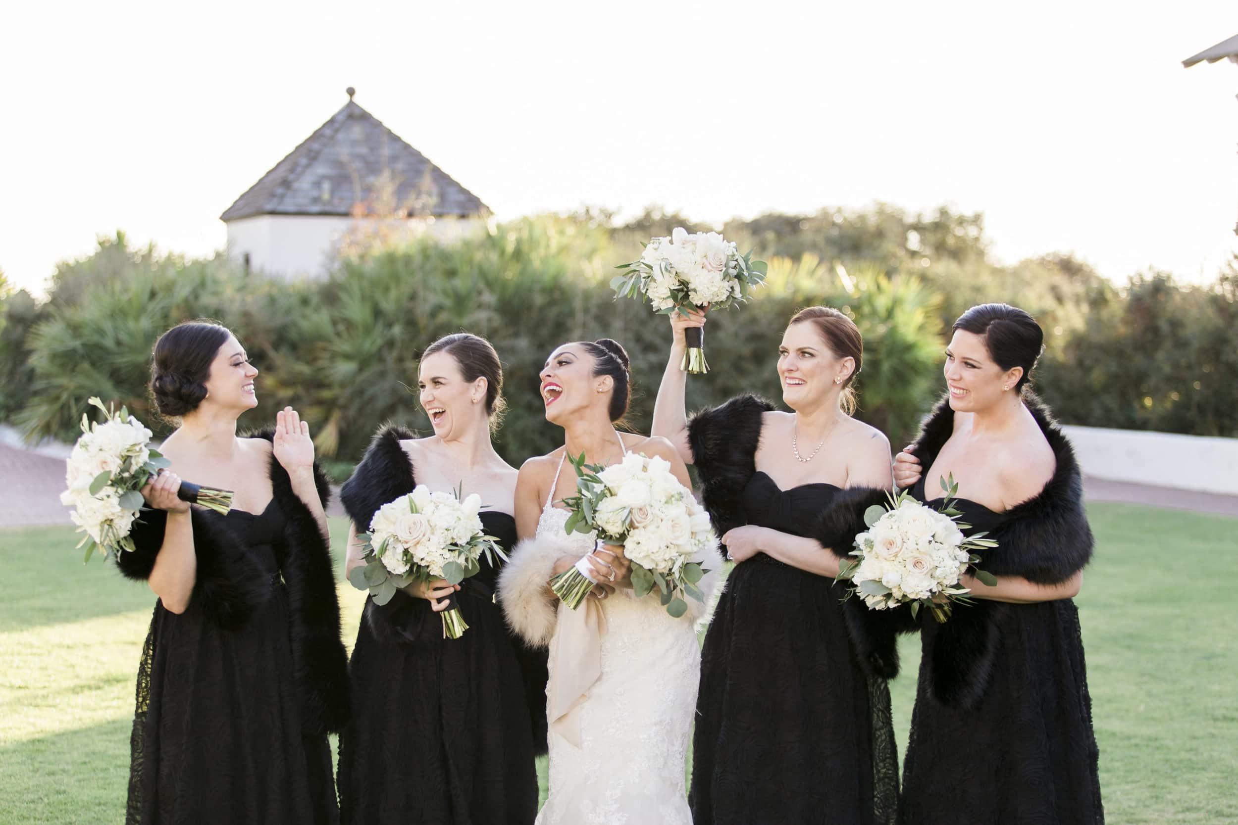 bridesmaids in all black dresses with black fur laughing with bride in all white