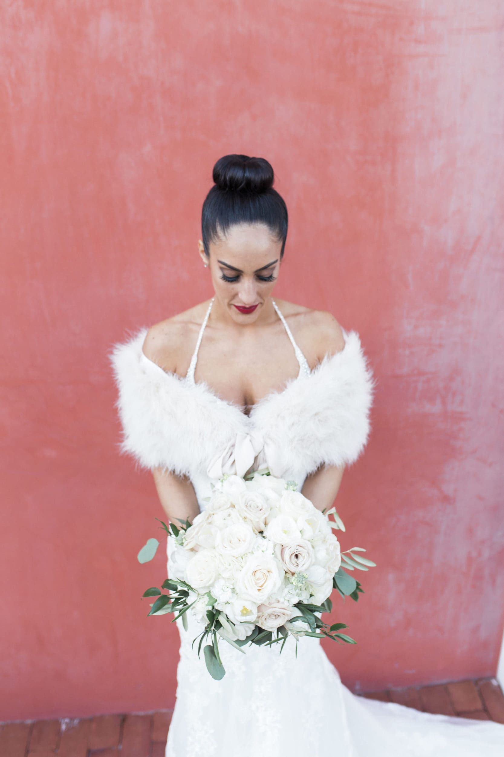 vogue bride in pronovias dress with high bun and red lips and white fur shawl in front of red wall
