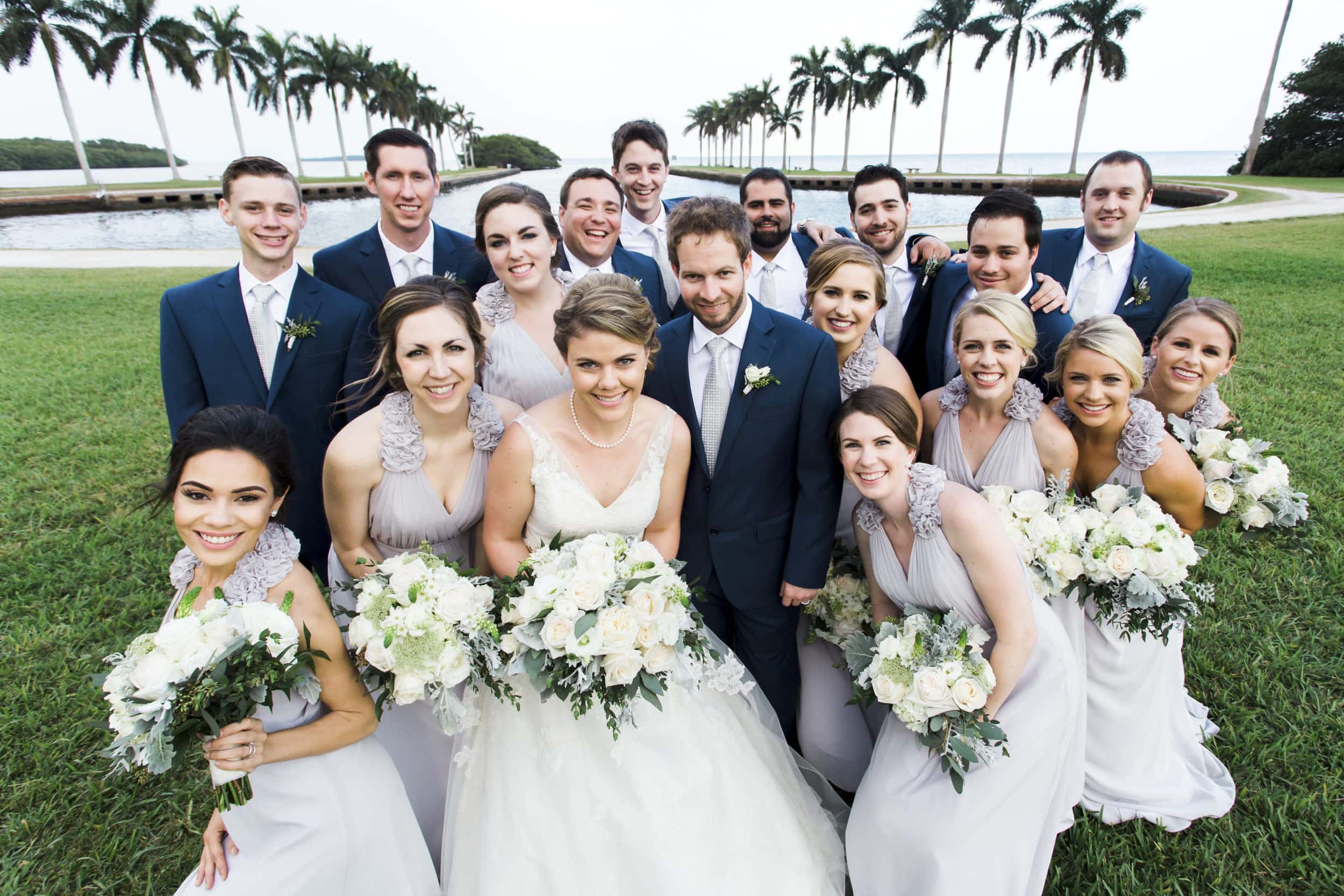 bride in white gown with groom in navy tuxedo with bridesmaids in grey flowy dresses and groomsmen in navy tux's with white flowers in front of palm tress and water at deering estate wedding