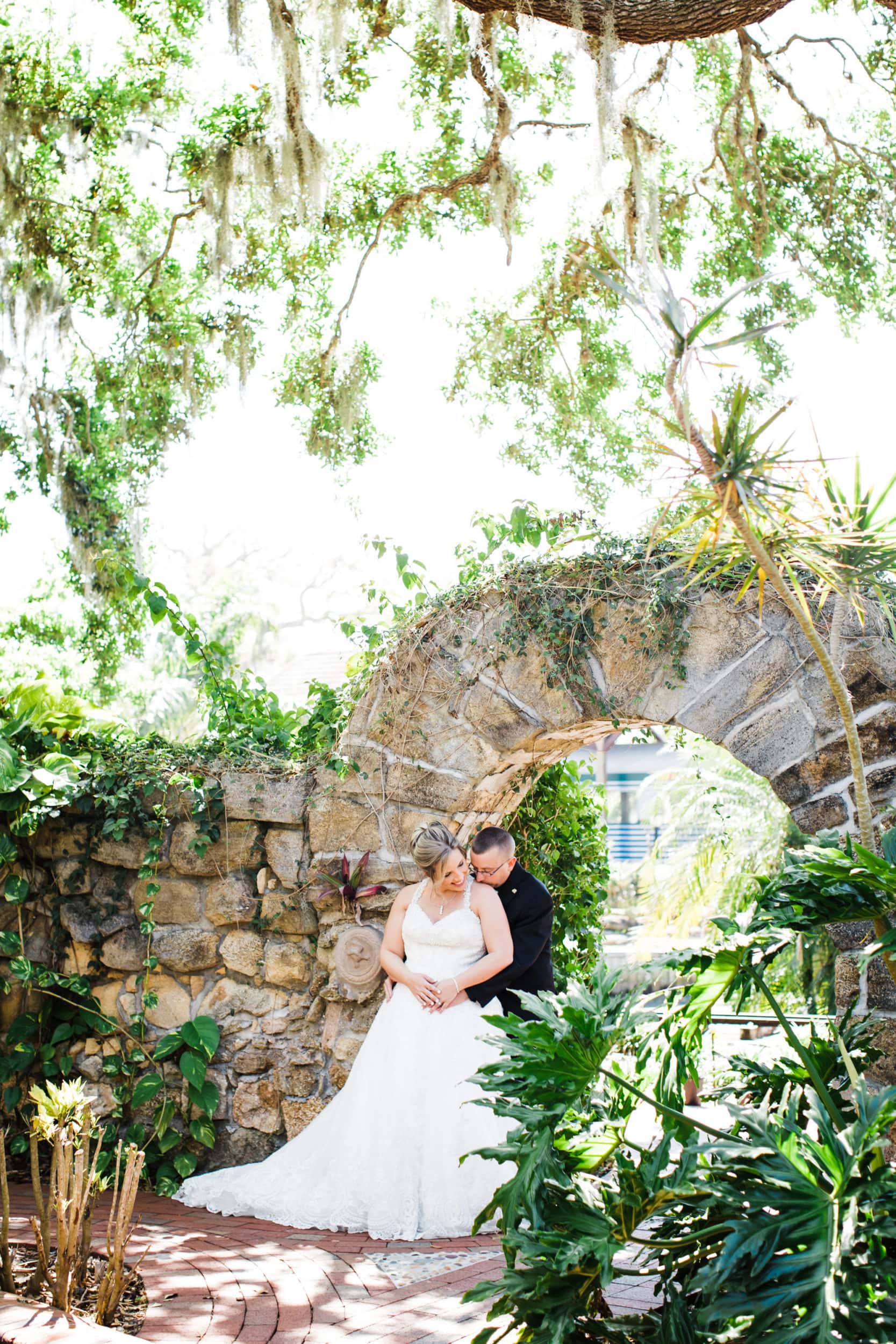 Groom hugging bride from behind in front of stone wall arch and walkway with lush green trees