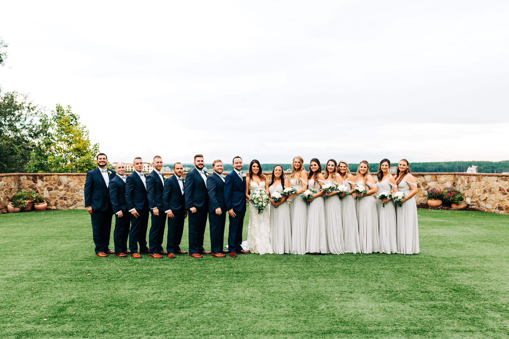 bridal party in grey dresses and groomsmen in black tuxes formal picture on front lawn of Bella Collina with bride and groom