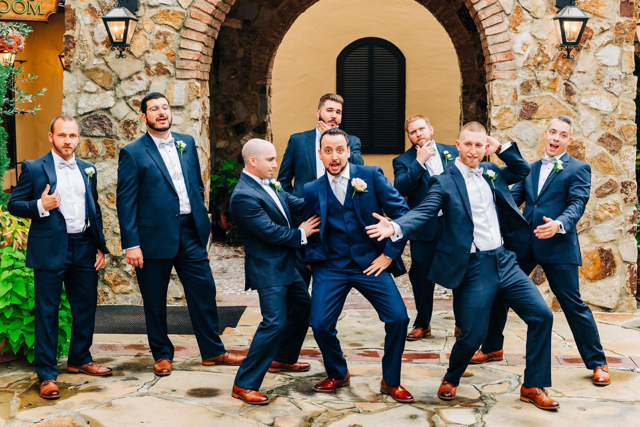 funny group shot of groom and groomsman in navy blue tuxes in front of stone archways 