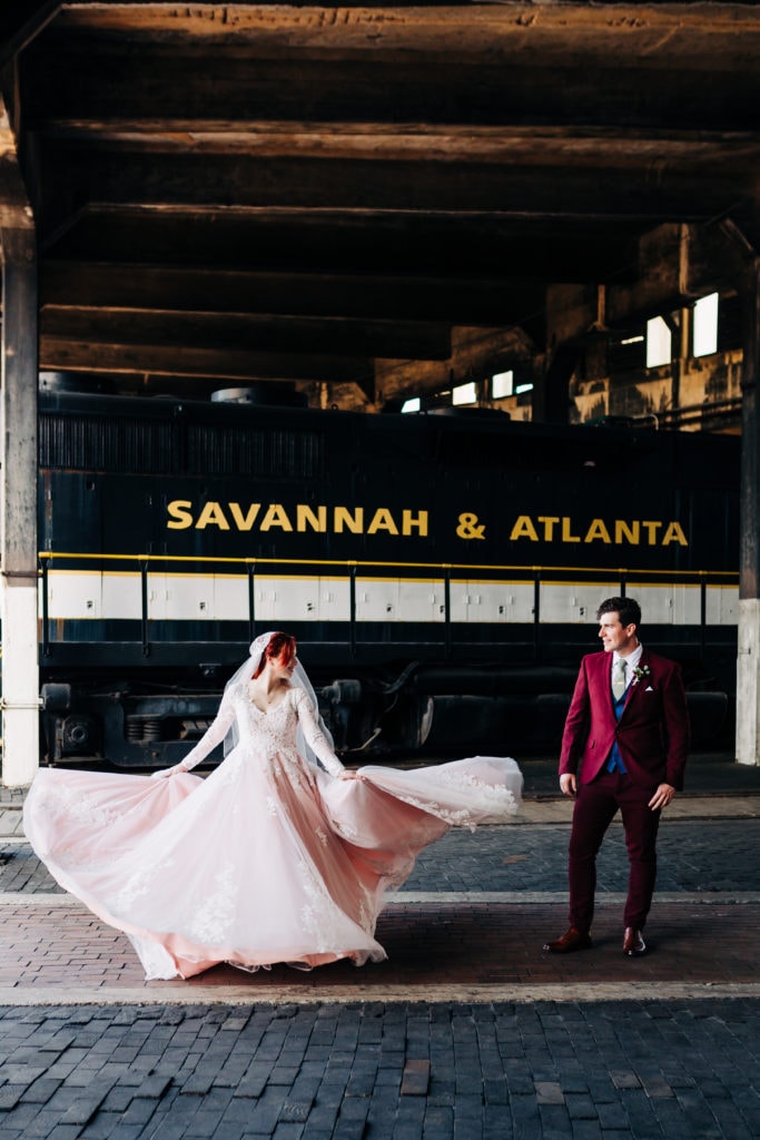 bride in long sleeved blush gown and cathedral veil spinning next to groom in custom fitted maroon suit at the Georgia state railroad museum in savannah