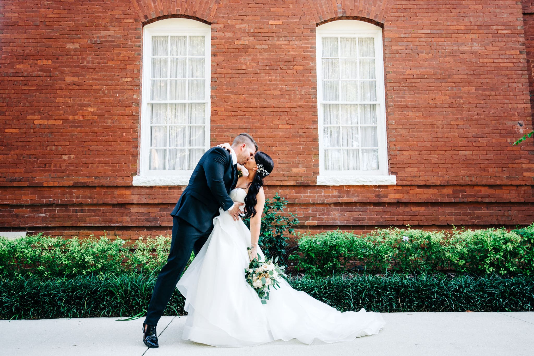 groom kissing and leaning back bride in front of red brick wall at venue 1902 in sanford