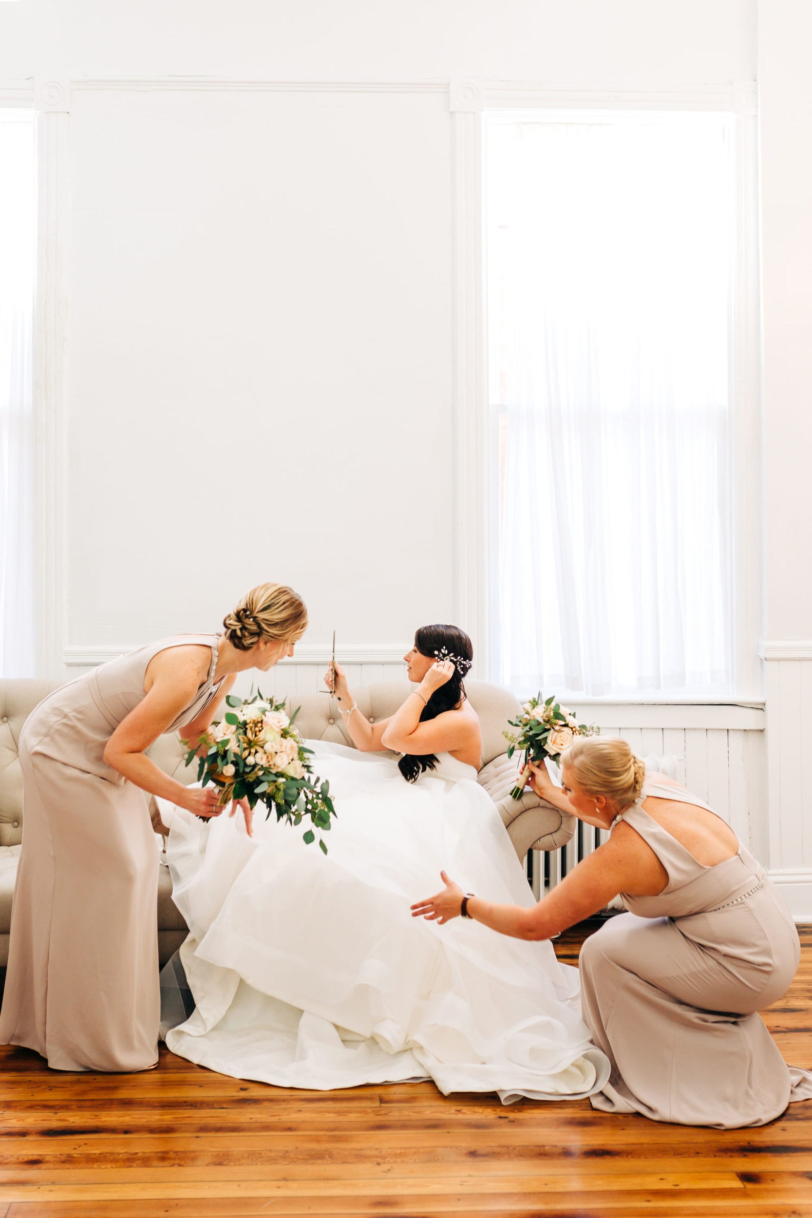 bride sitting on couch getting ready with two bridesmaids fluffing her dress