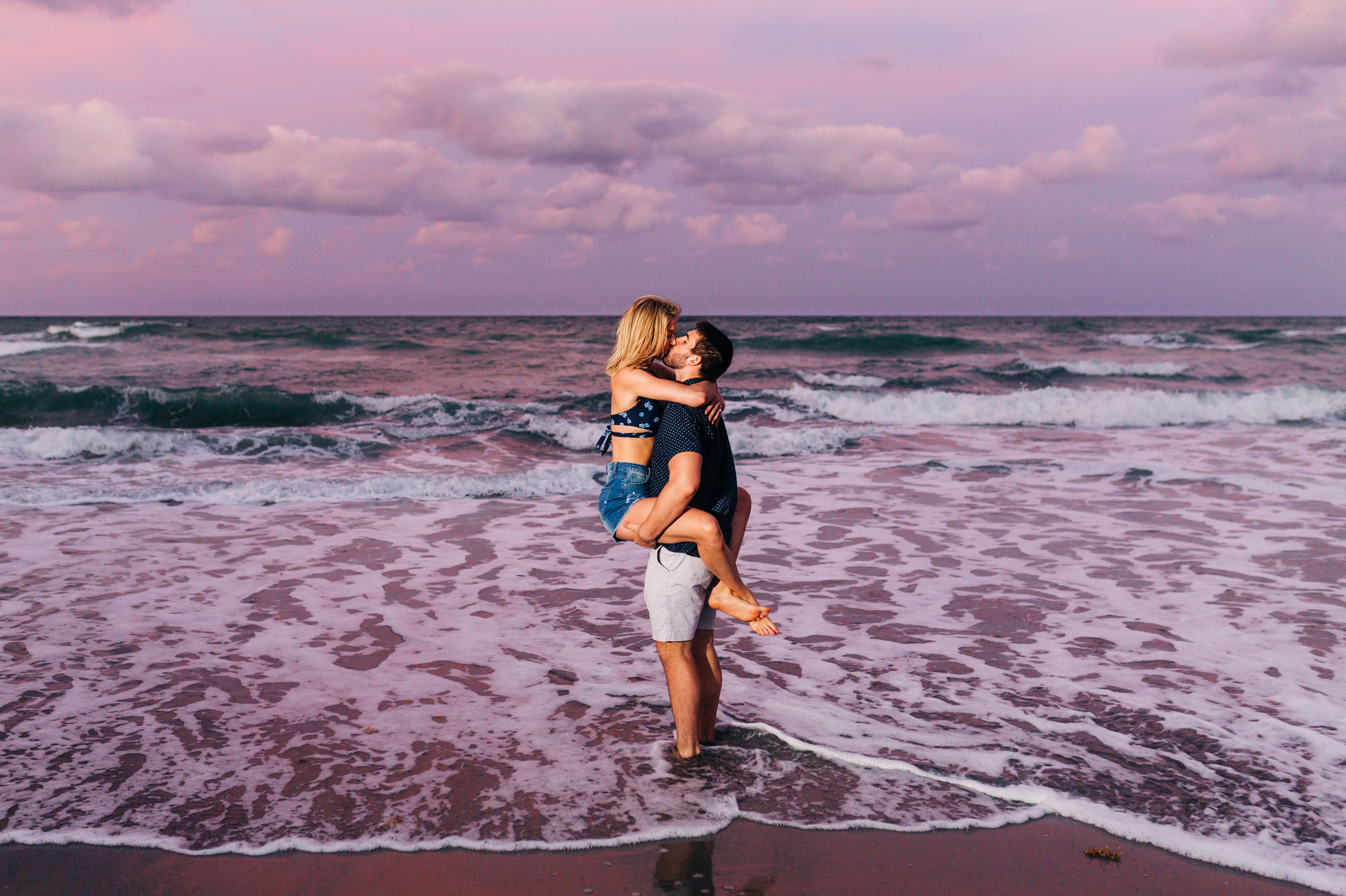 girl wrapped around boy's hips standing in the ocean with purple sunset behind them at blowing rocks preserve