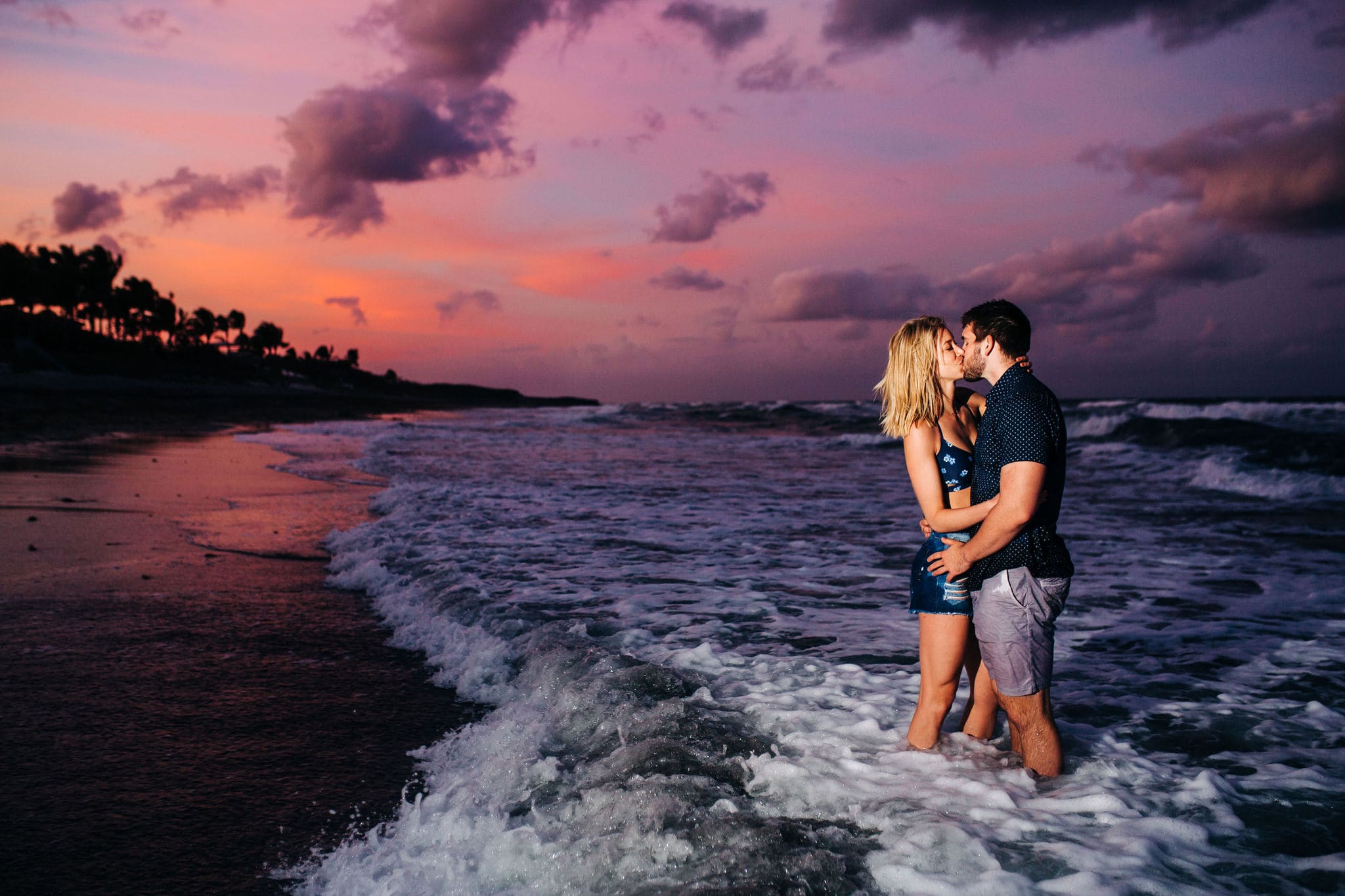 couple kissing in the ocean with a pink and purple sunset behind them at blowing rocks preserve