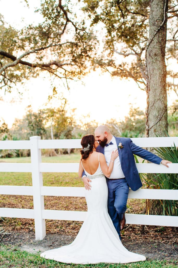 bride with a low back wedding dress kissing groom in custom navy suit with American flag details at up the creek farms wedding venue along white fence with sunset in background