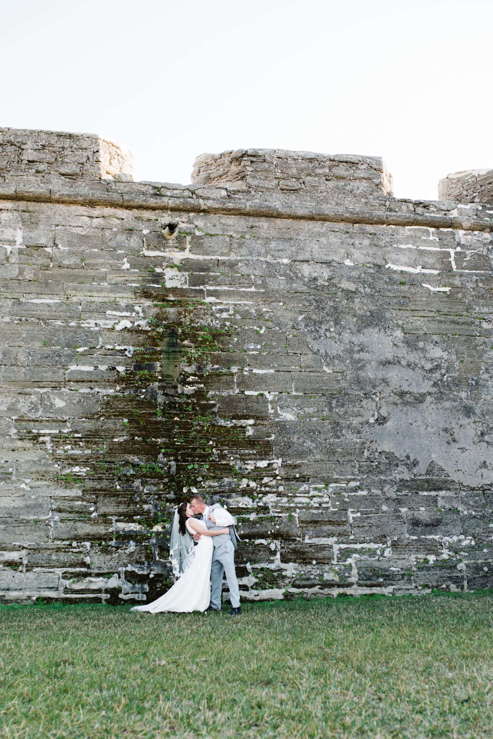bride in white dress with groom in grey suit with large stone castle wall with green lawn at castillo de san marcos wedding