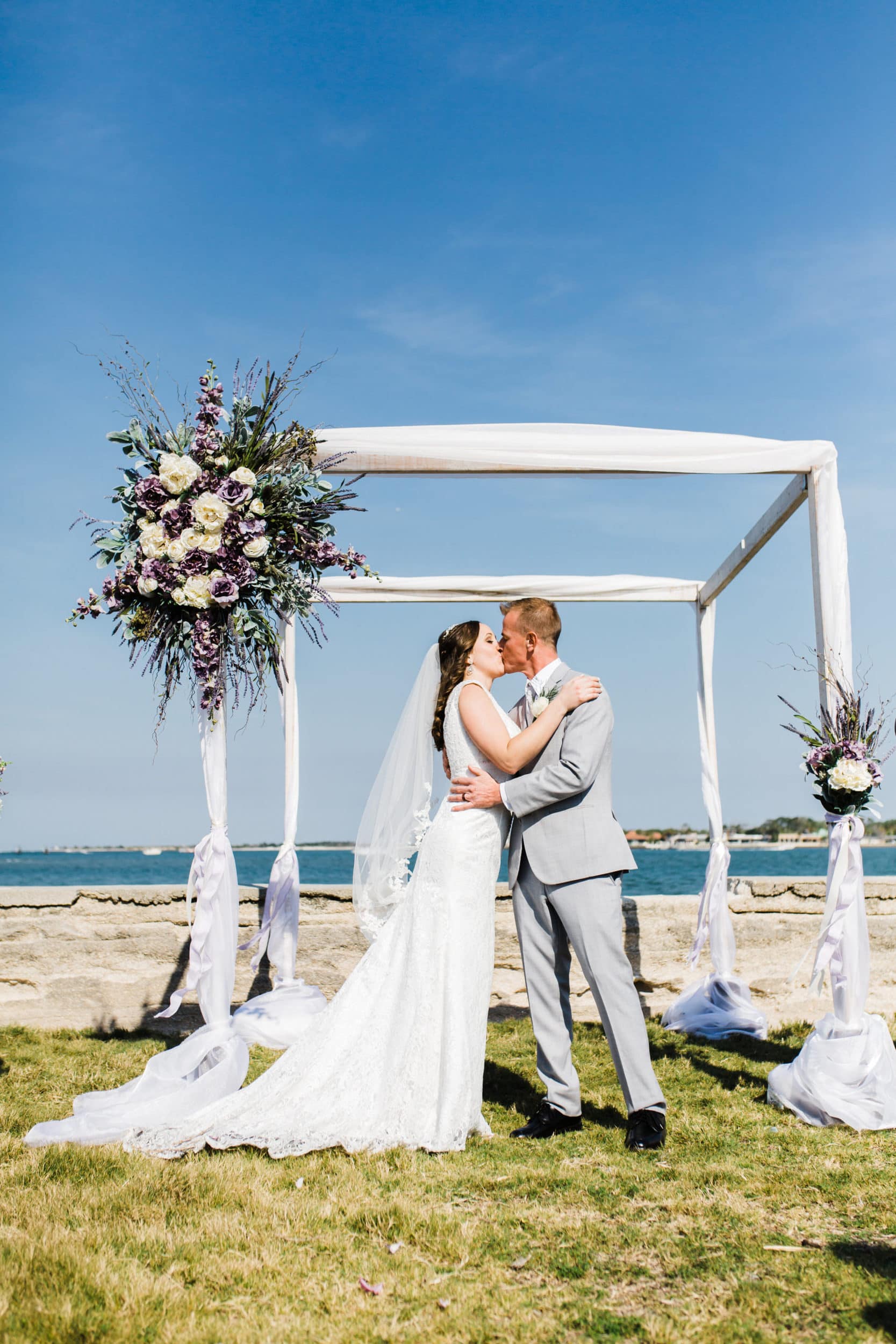 bride with white dress and groom with grey suit with large altar with white drapery with white and lavender flowers with ocean view at castillo de san marcos