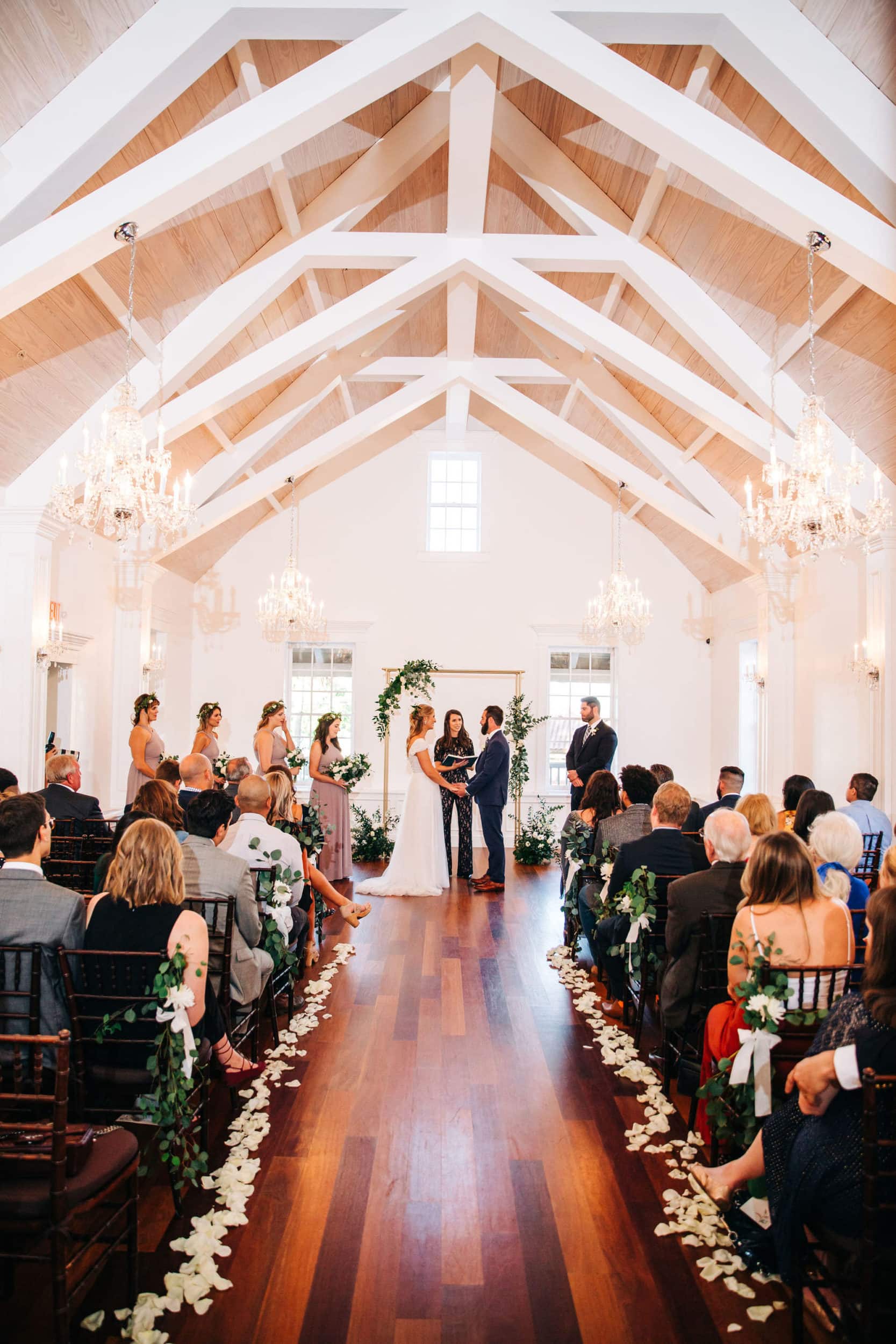 bride and groom with gold arch with greenery and white flower garland with cathedral ceiling with aisle and lined up chairs at white room wedding
