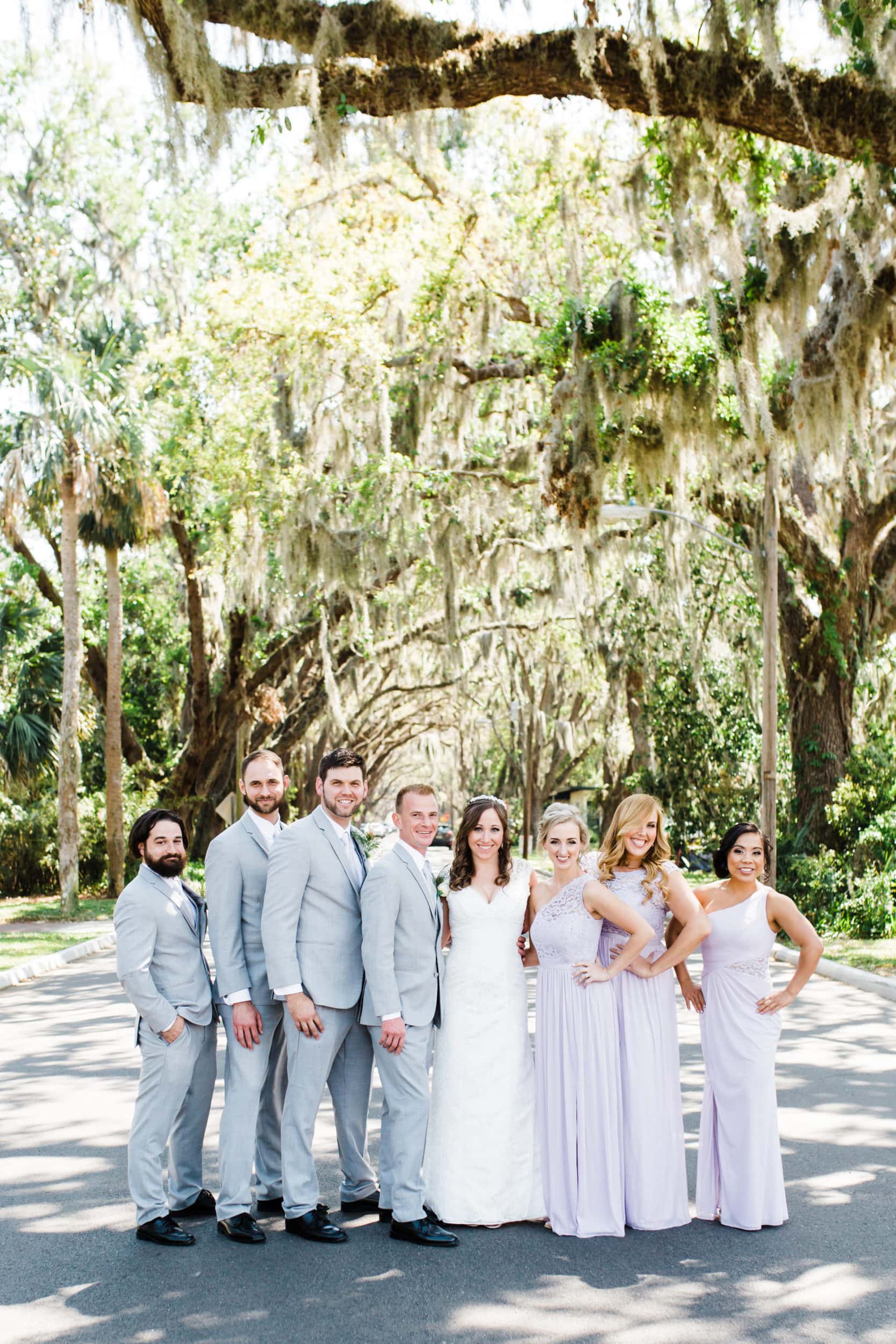 bride and groom with bridesmaids with lavender dresses with groomsmen with grey suits with large road with large oak tress with spanish moss at castillo de san marcos wedding
