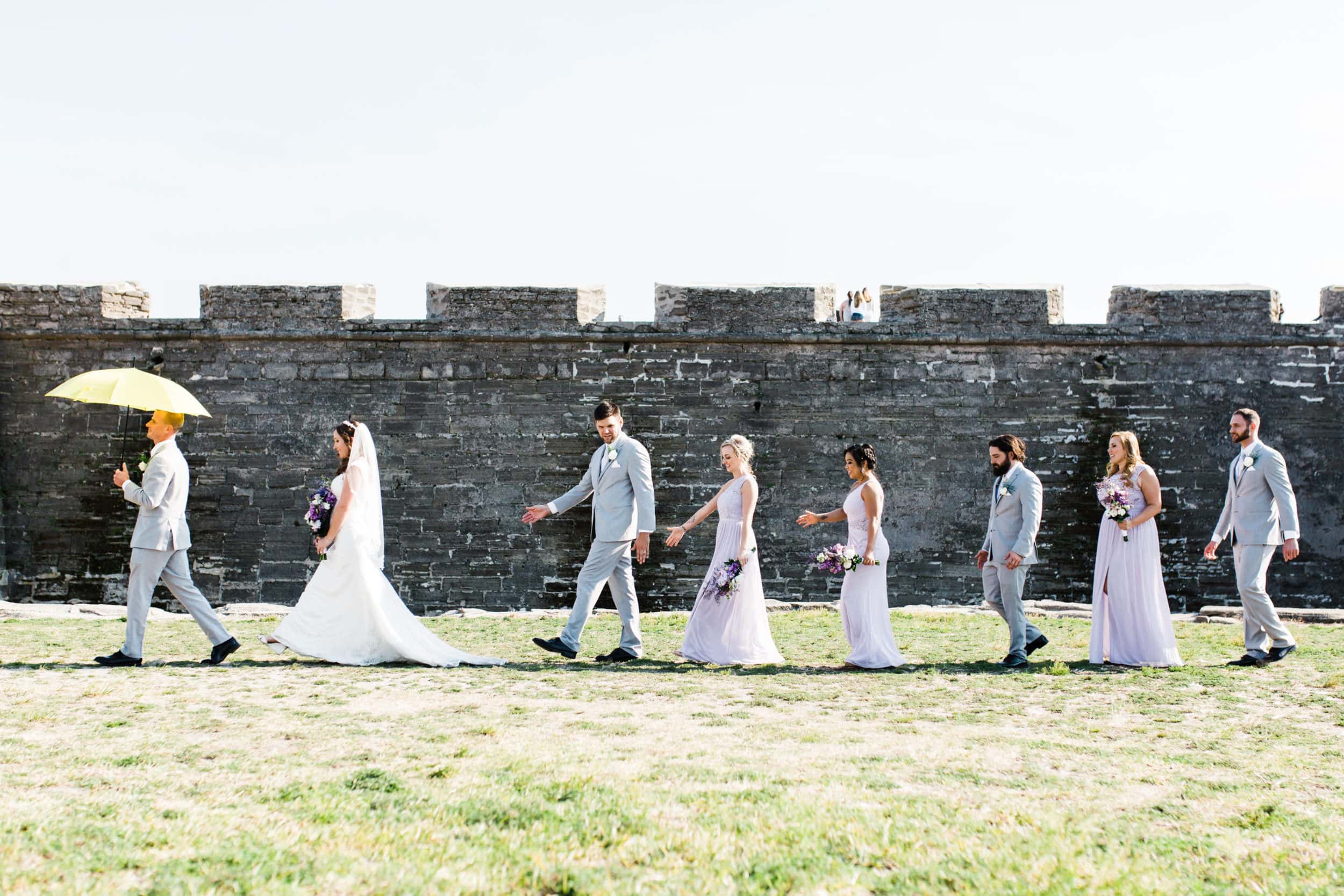 bride and groom with bridesmaids and groomsmen in line with yellow umbrella with grey suits with lavender dresses with large castle stone wall at castillo de san marcos wedding