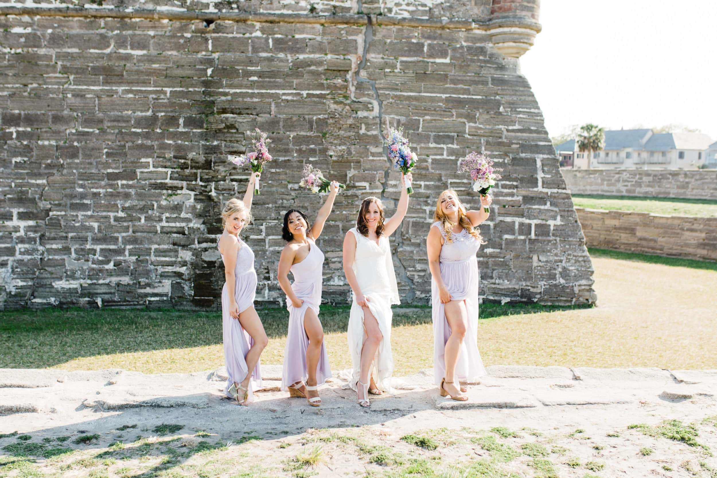 bridesmaids with white and purple flowers with lavender dresses with large stone wall in background at castillo de san marcos wedding