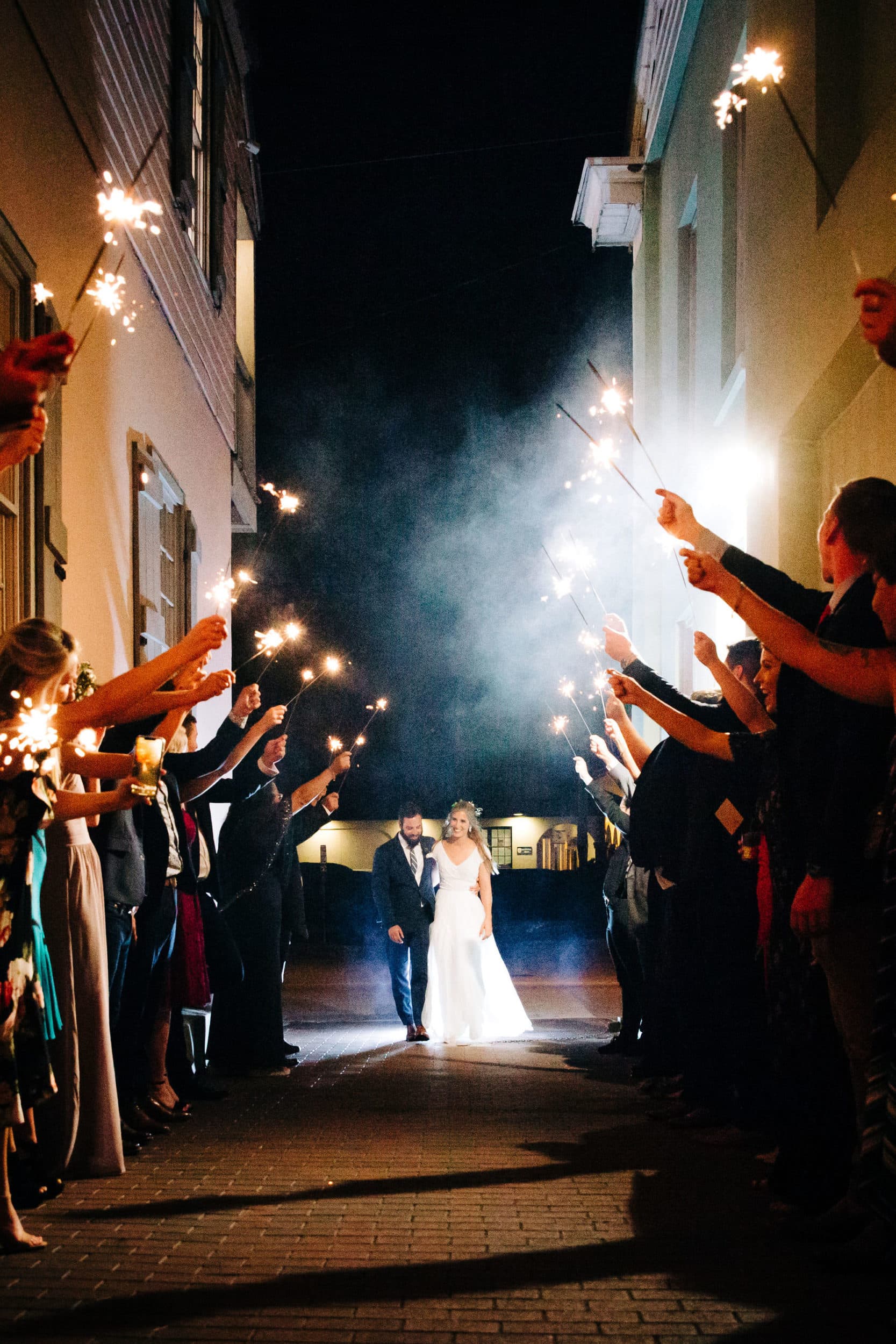 bride and groom with lined up crowd holding sparklers in alleyway at white room wedding
