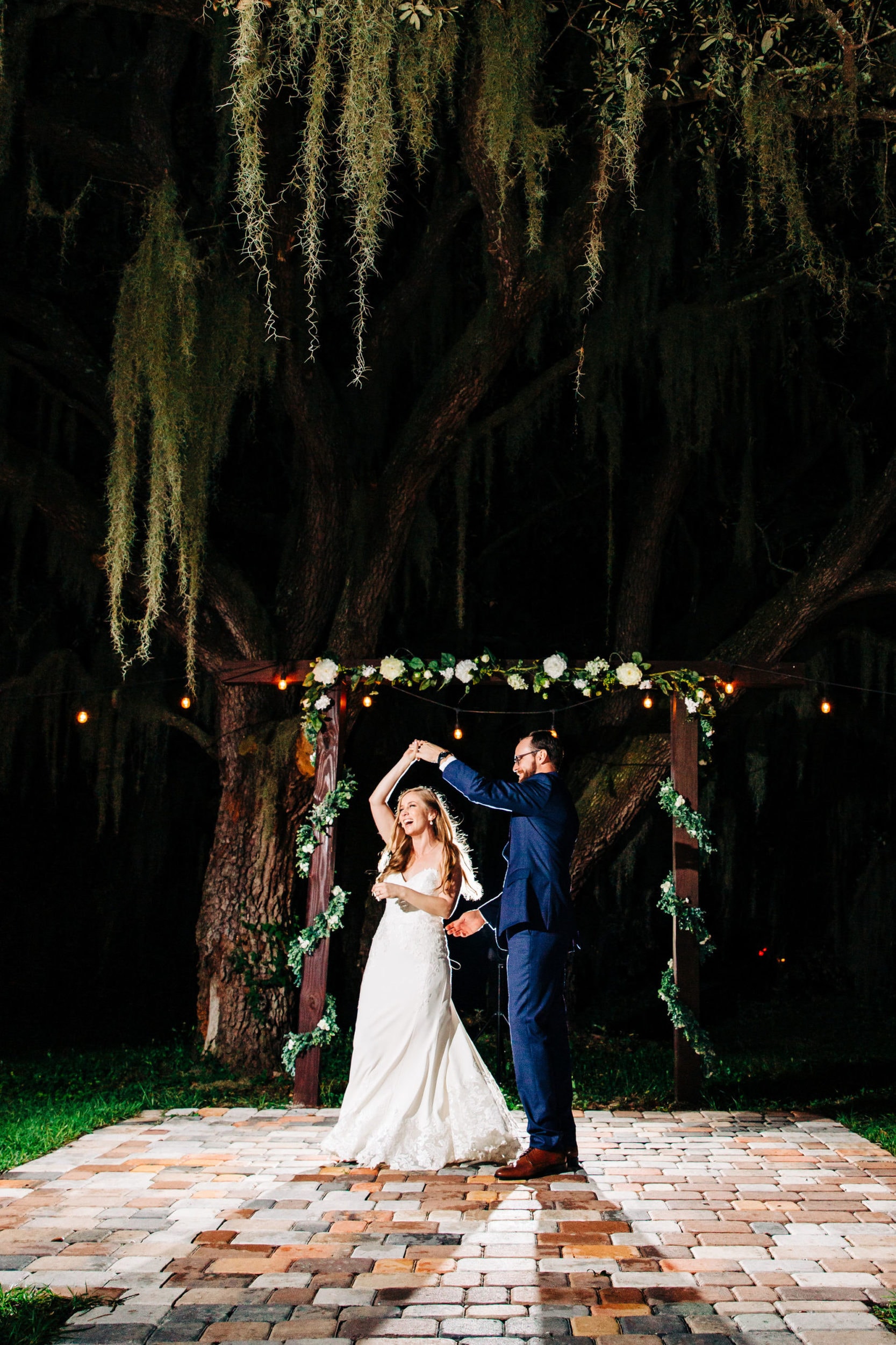 bride in white gown and groom in navy tuxedo in front of large oak tree with wooden arch with white flower garland at ever after farms ranch