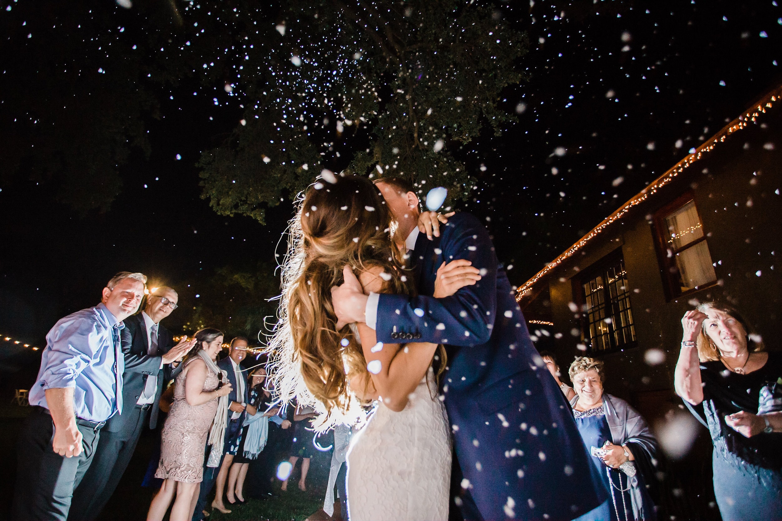 bride with white dress with groom with navy suit with white confetti with cheering wedding guests in background at the acre wedding