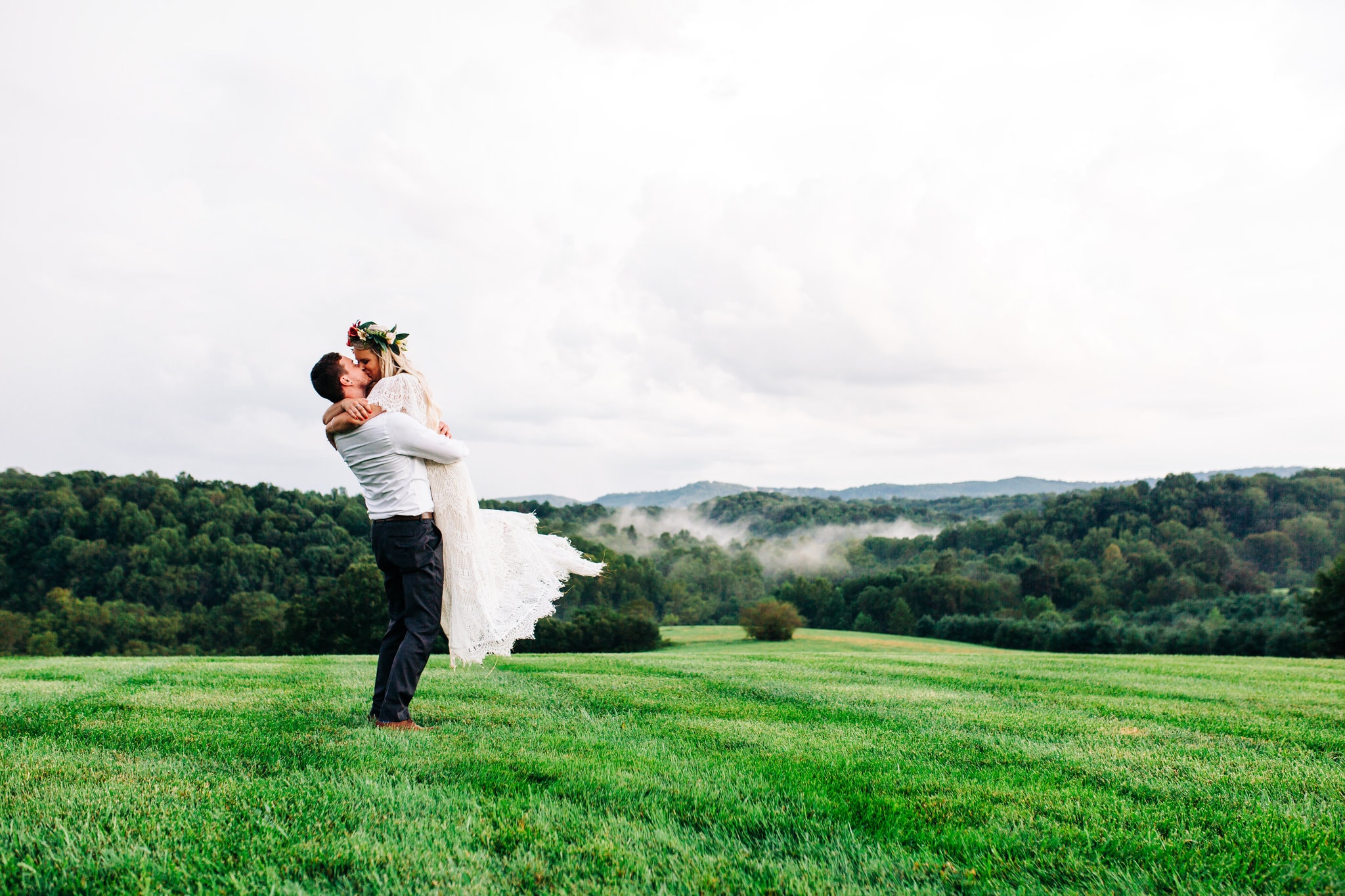 groom with grey and white suit holding up bride with white lace dress with flower crown with foggy mountain views in background at Boho Gambill Estate Wedding