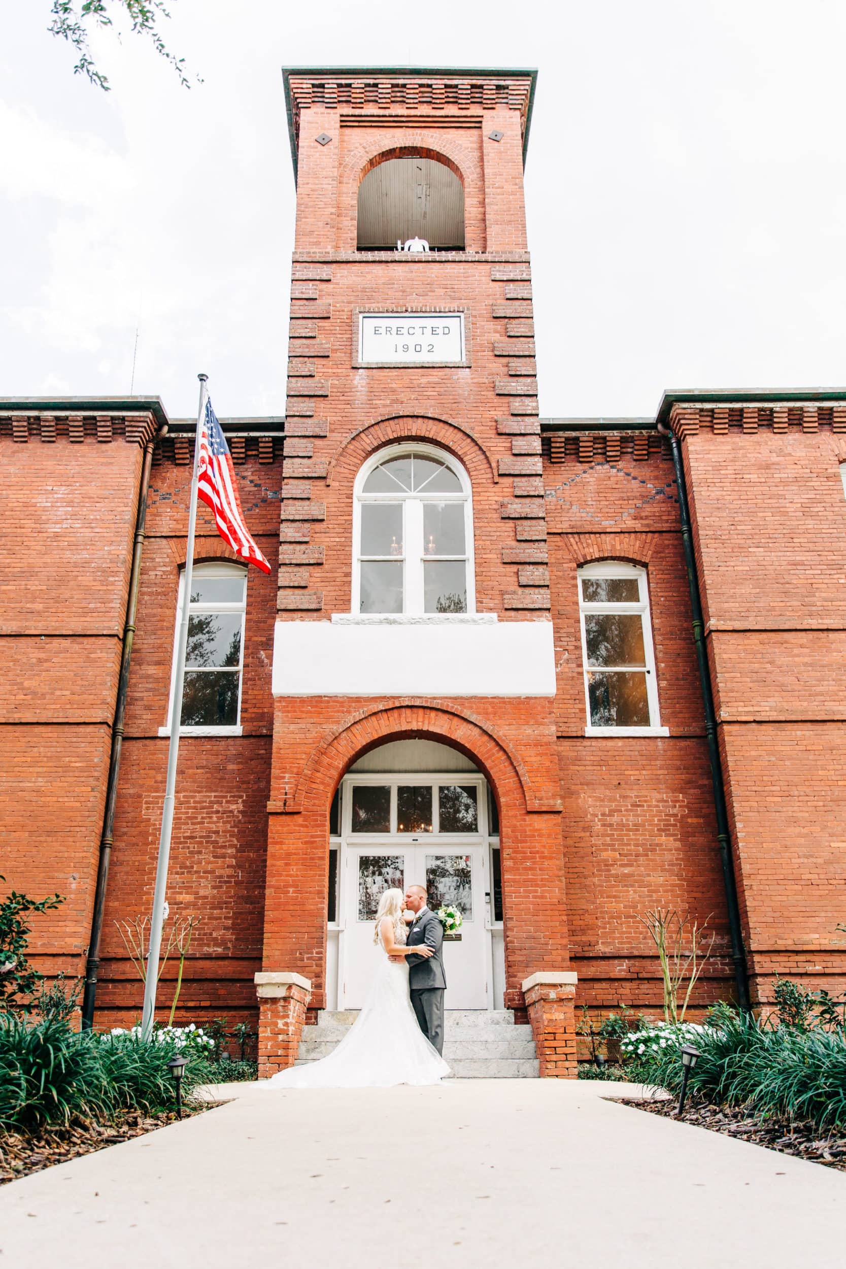bride in white dress and groom in grey suit with large brick building with white french door entrance at venue 1902 wedding