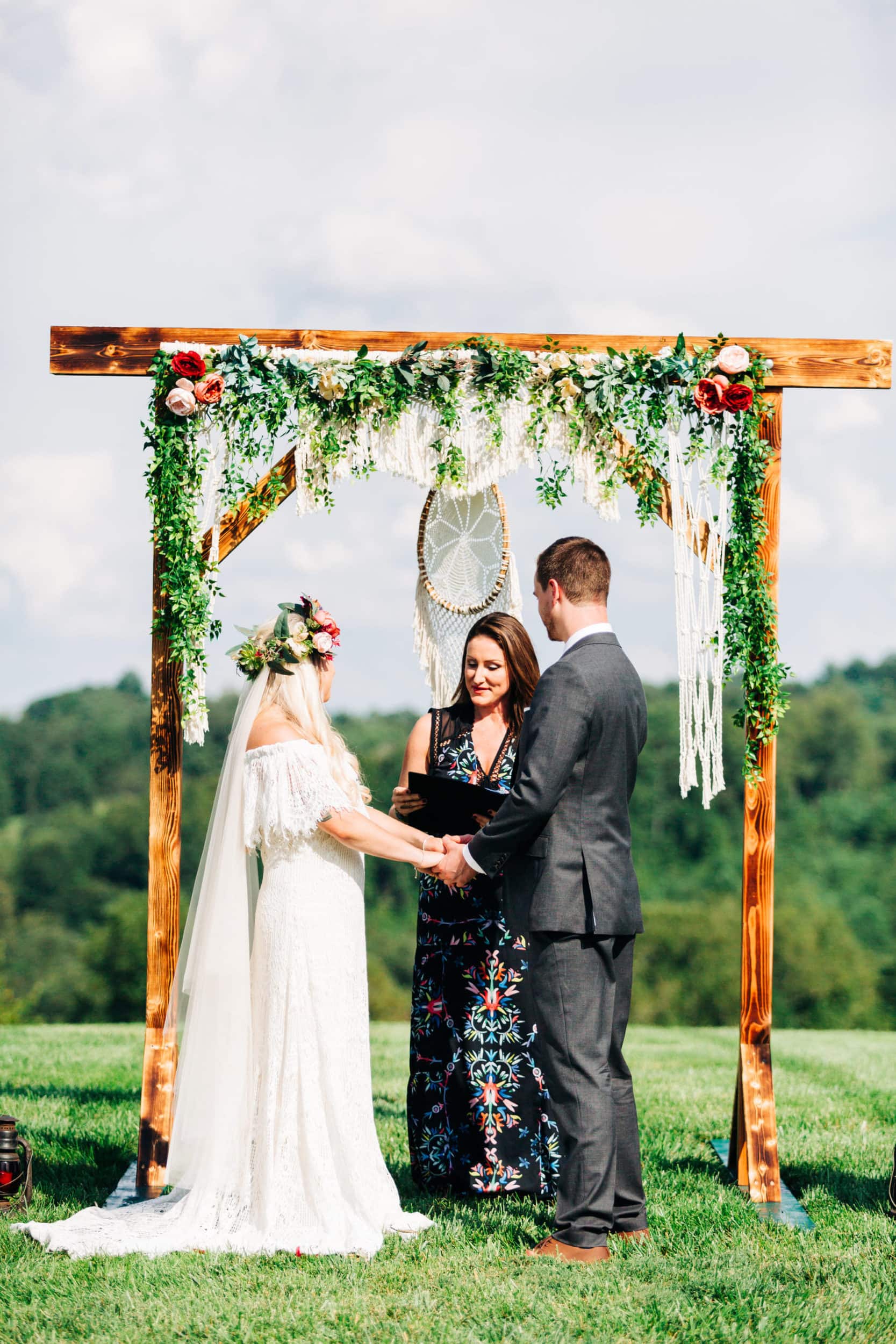 bride with lace off the shoulder dress with groom with grey suit with wood archway with greenery garland with white macrame in background at Boho Gambill Estate Wedding