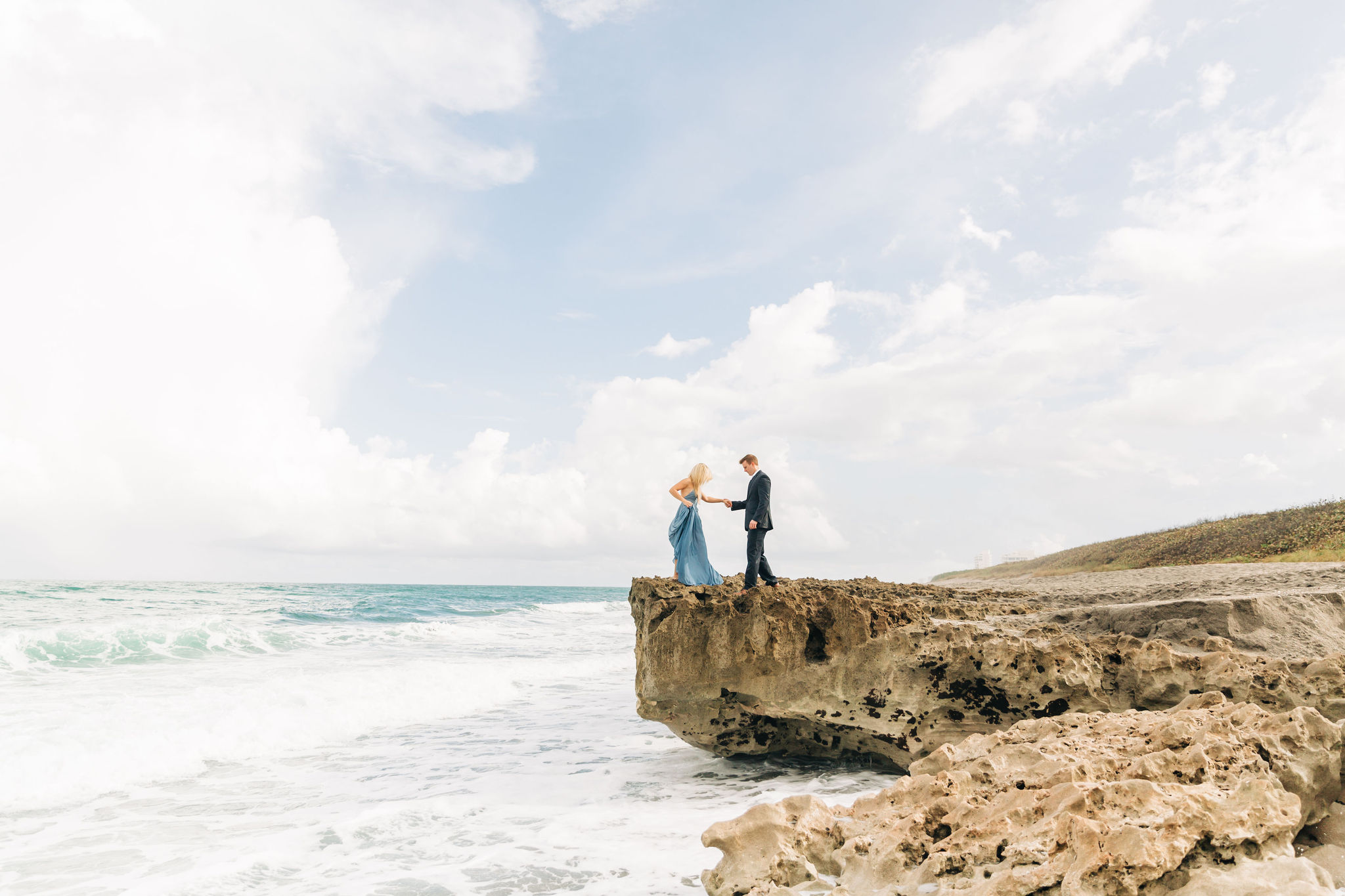 girl with blue dress holding hands with boy with blue suit on rocky beach at Orlando Engagement Session Locations