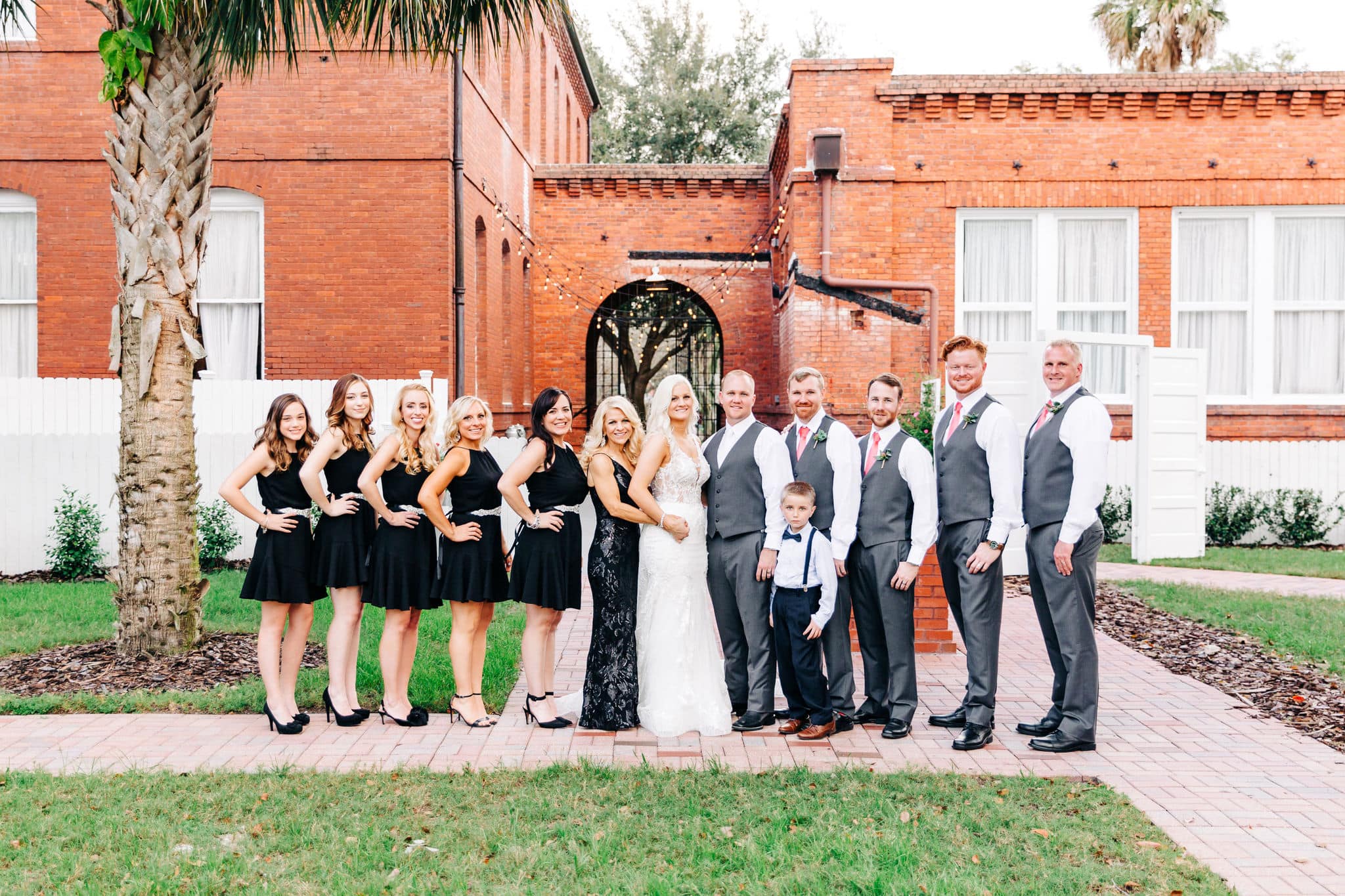 bride and groom with bridesmaids with black dresses with groomsmen with grey suits with large red brick building in background at Venue 1902 Wedding