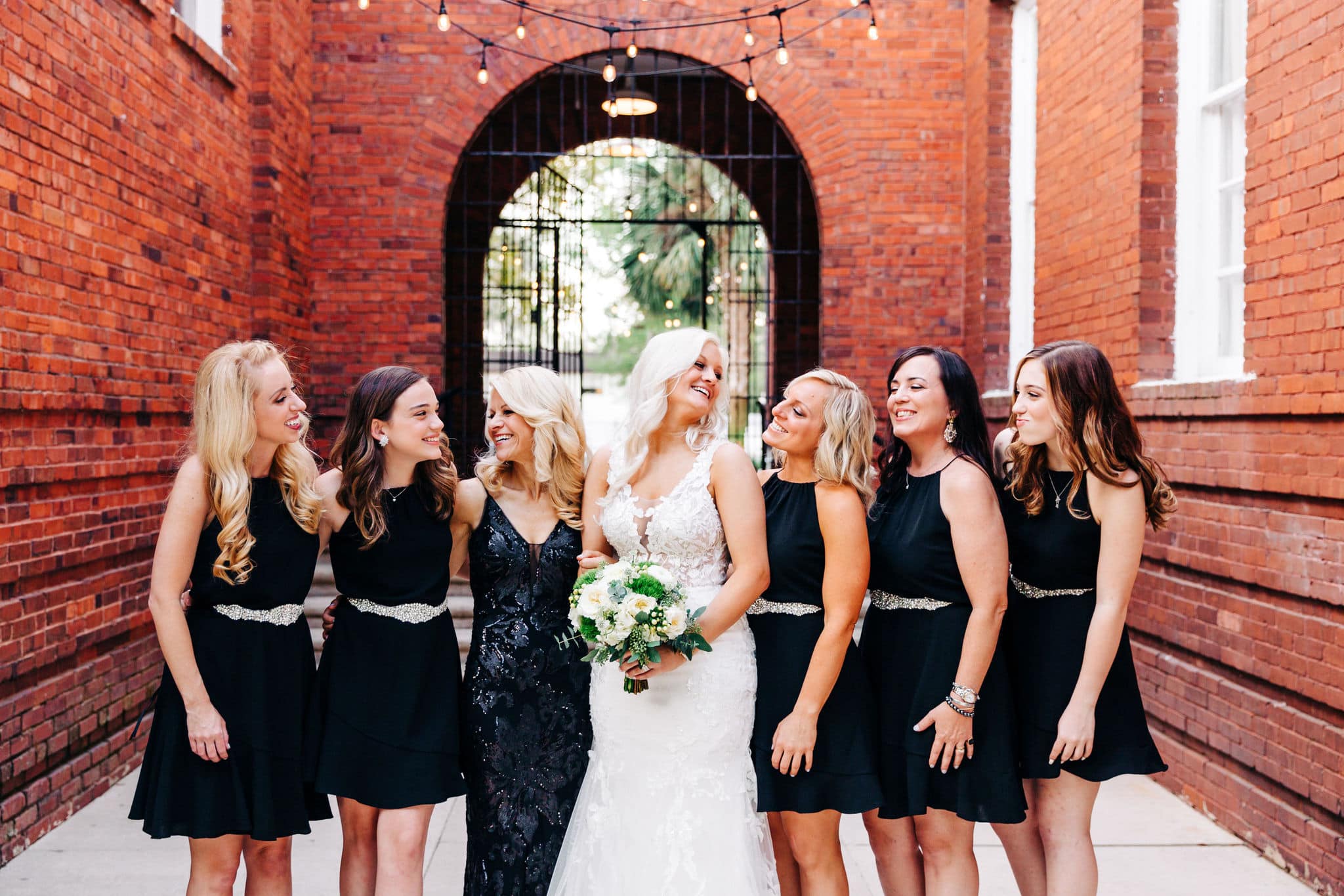 bride in white dress with white bouquet with bridesmaids with black dresses with brick building with wrought iron gate in background at Venue 1902 Wedding
