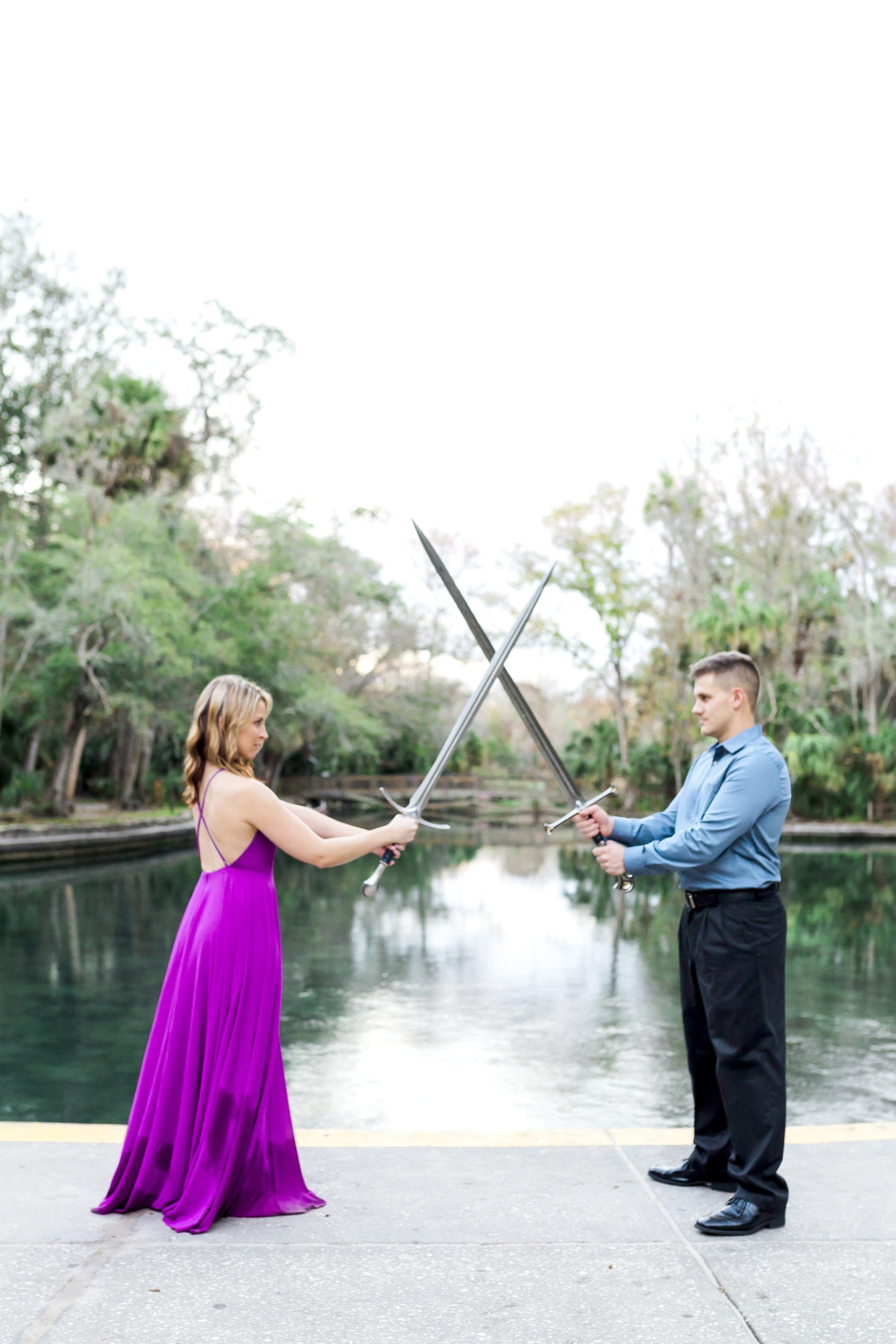 bride to be with flowy purple dress with groom to be with swords posing with pond in background