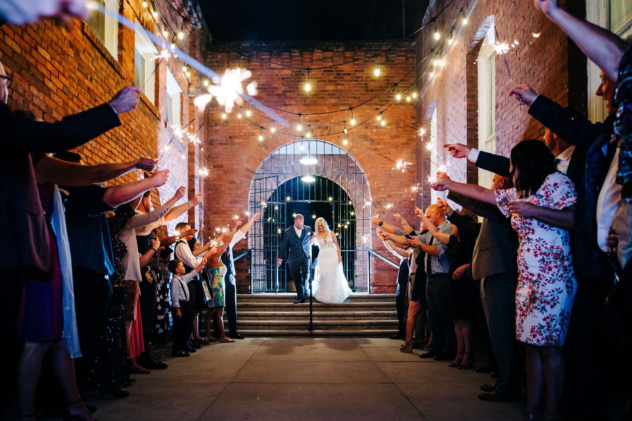 bride with white dress with groom with black suit with wedding guest crowd with sparklers with large brick building with wrought iron gate in background at Venue 1902 Wedding