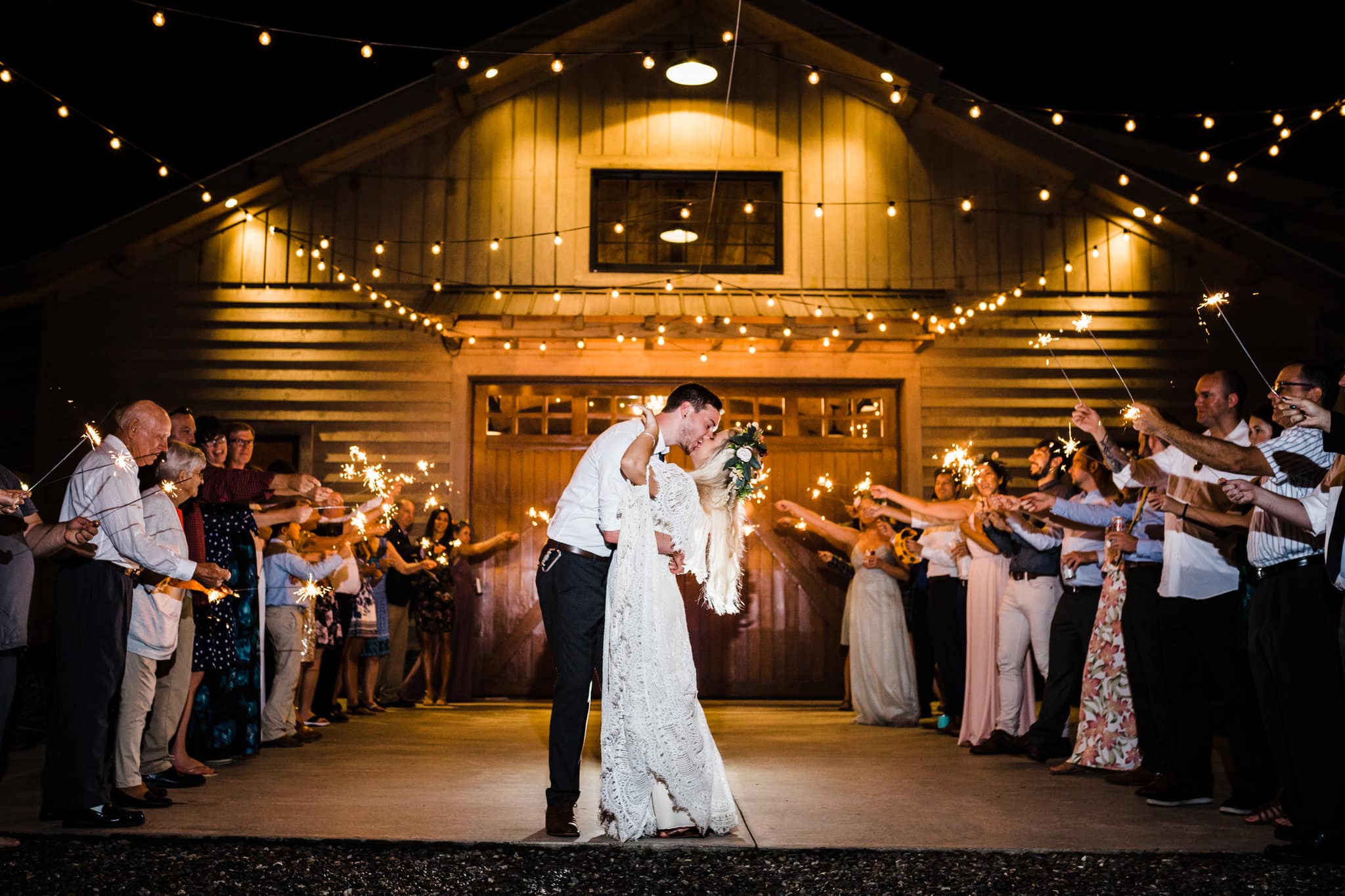 bride and groom kissing between wedding guests with sparklers with large barn with string lights in background at Boho Gambill Estate Wedding