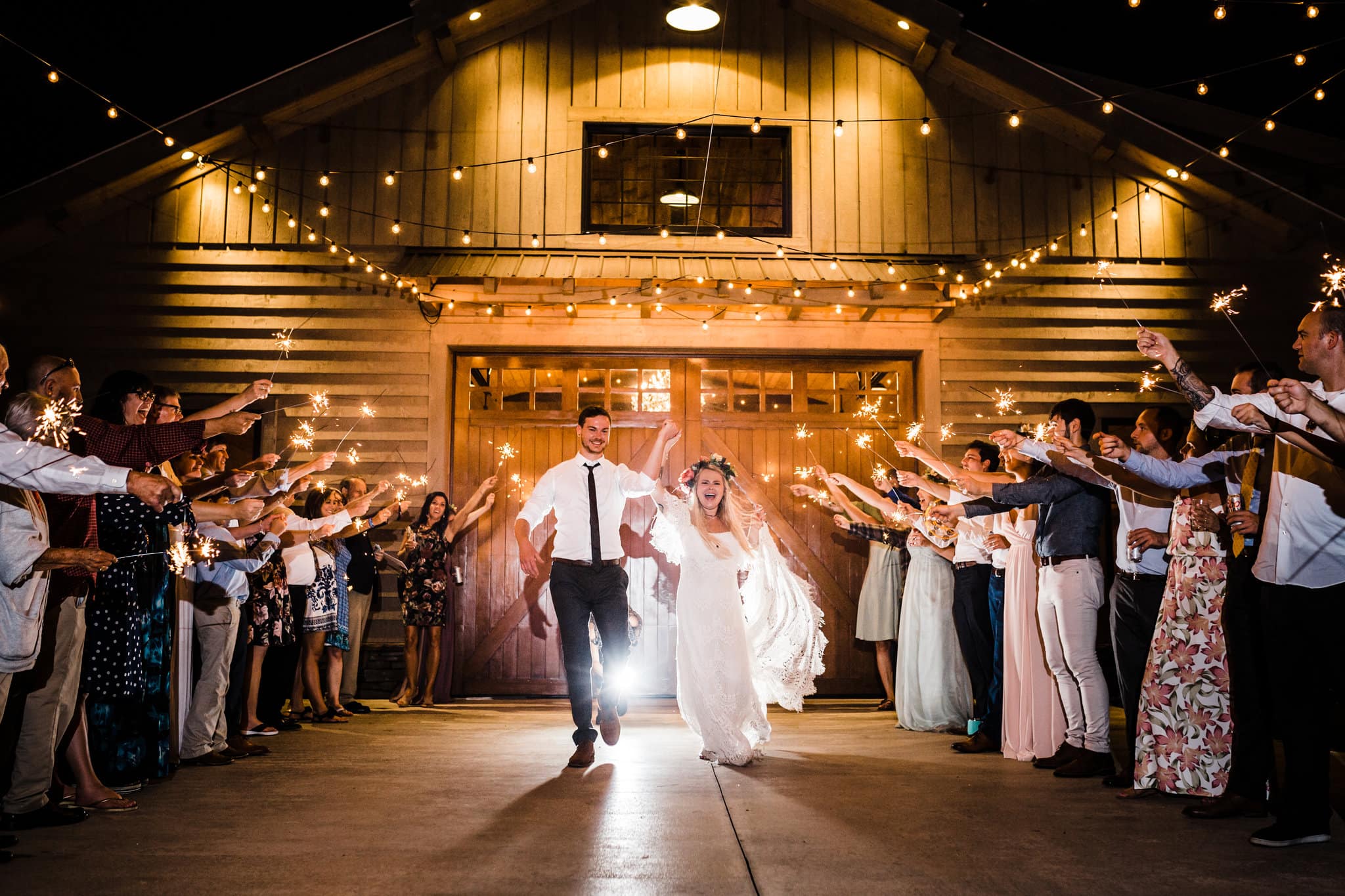 bride and groom walking between wedding guests with sprinklers with large barn with string lights in background at Boho Gambill Estate Wedding