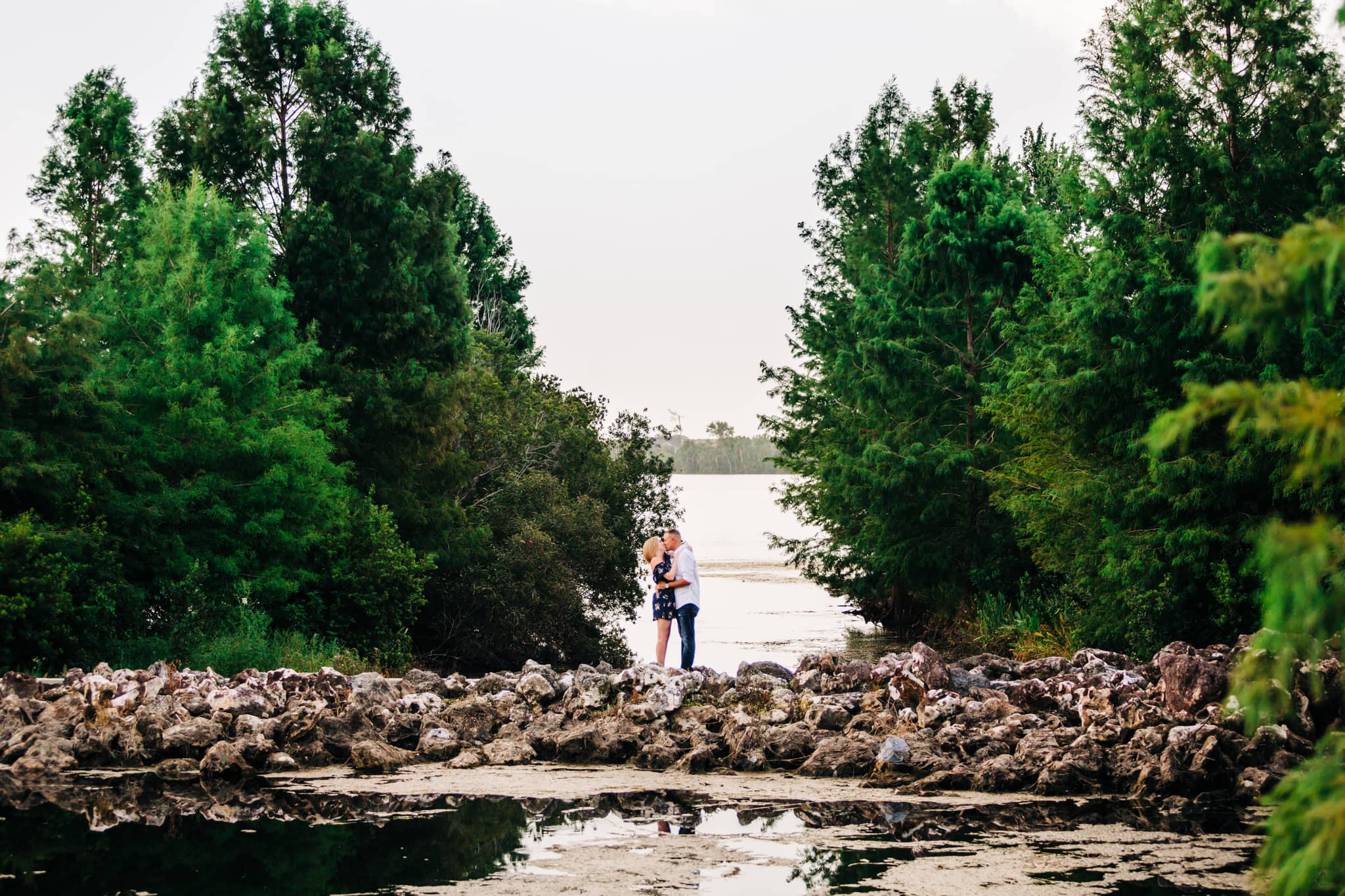 bride to be and groom to be with large rocks with lake water with large trees in background